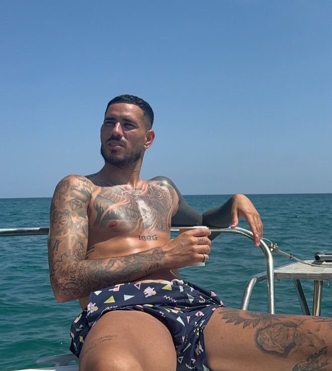 Jogador Tonny Sanabria sem camisa, repleto de tatuagens, relaxando em um barco no meio do mar sob céu aberto.