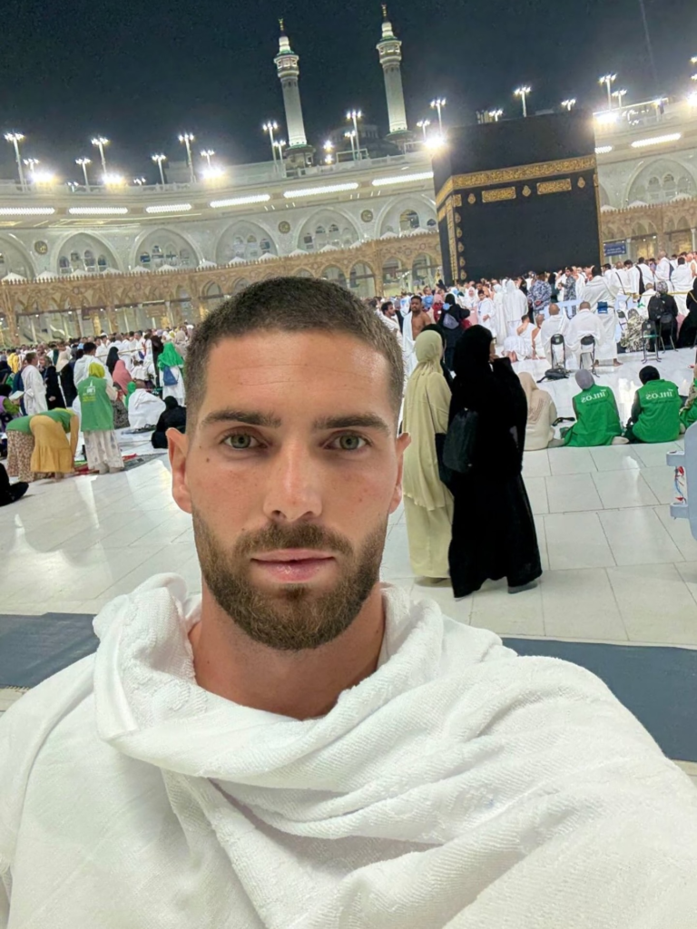 Jogador Luca Zidane usando trajes tradicionais Ihram em selfie com a Caaba ao fundo, durante visita à Meca.