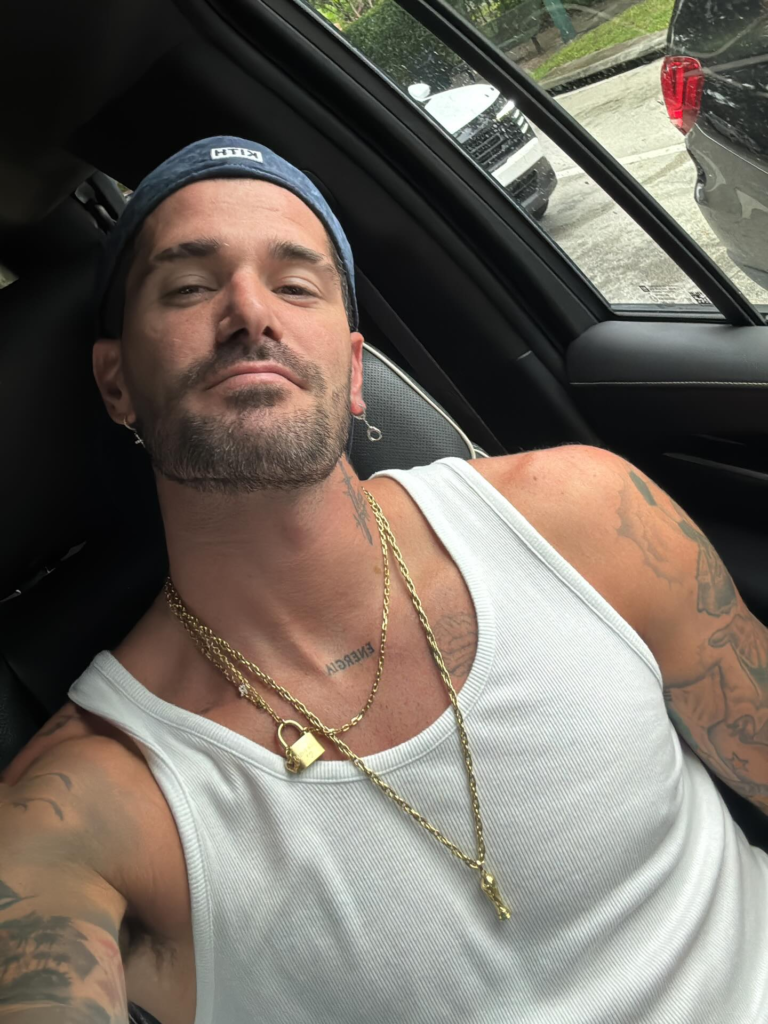 Rodrigo de Paul posando para selfie dentro de um carro, usando regata branca, boné azul da marca KITH, correntes douradas e exibindo tatuagens no pescoço e braços.