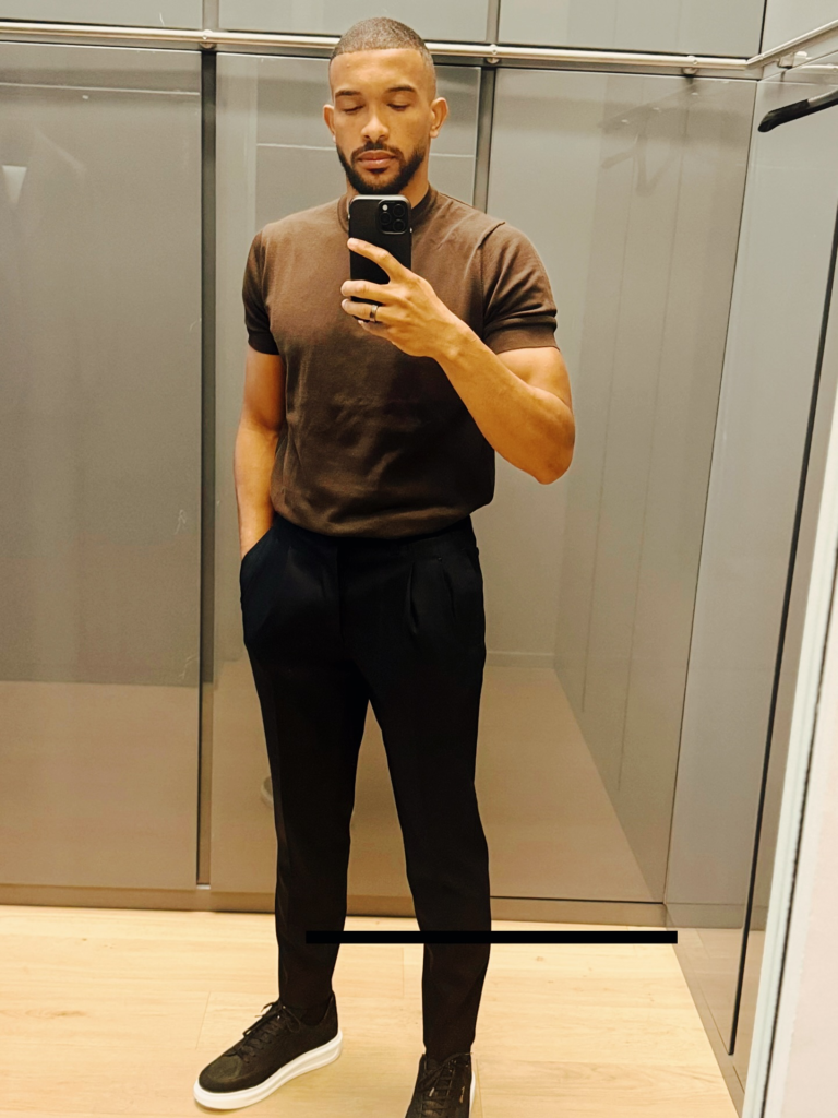 Bremer posando para foto em frente ao espelho, vestindo camiseta marrom, calça preta e tênis casual com solado plataforma branco.