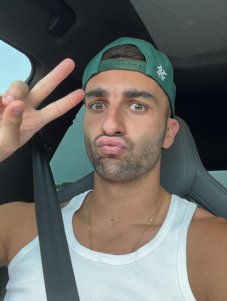 Ferran Torres tirando uma selfie de 'duck face' dentro de um carro, fazendo o sinal da paz com os dedos, usando um boné verde virado para trás, uma regata canelada branca e uma corrente dourada.
