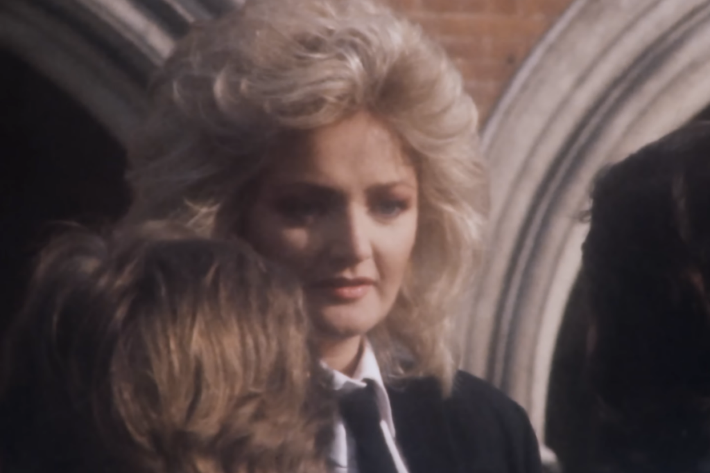 A cantora Bonnie Tyler, com seu característico cabelo loiro volumoso dos anos 80, olha para a câmera com uma expressão séria, vestindo uma camisa branca e gravata preta sob um blazer. Ela está em primeiro plano, com a parte de trás da cabeça de outra pessoa visível na frente e um arco de pedra ao fundo.