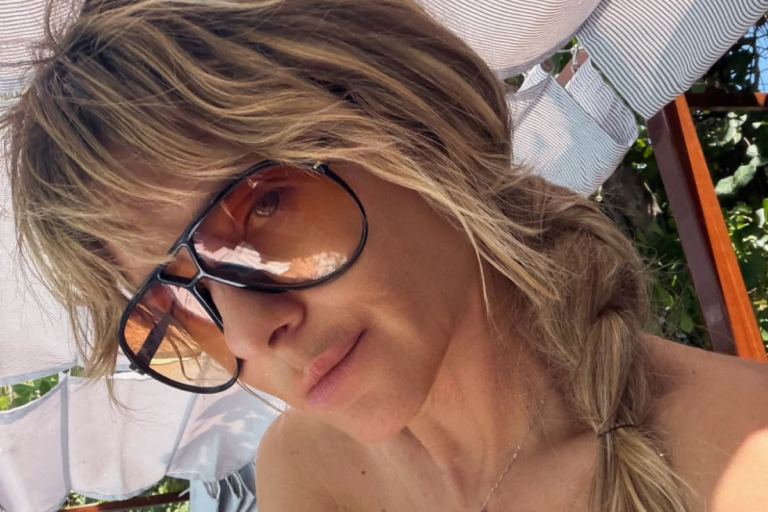 A atriz Carolina Dieckmann posa para uma selfie sob a sombra, usando óculos de sol grandes com lentes alaranjadas e o cabelo loiro preso em uma trança lateral.