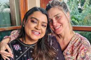 Preta Gil e Carolina Dieckmann sorriem abraçadas em uma varanda. Preta, à esquerda, veste blusa preta com bordados florais coloridos. Carolina, à direita com o braço sobre o ombro de Preta, veste blusa estampada em tons de rosa e bege. Ao fundo, detalhes de uma janela de madeira e folhagens verdes.