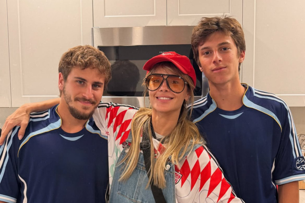 A atriz Carolina Dieckmann posa sorridente no centro, abraçada aos seus dois filhos, Davi e José, em um ambiente de cozinha. Ela usa um boné vermelho e óculos de sol, enquanto os filhos vestem camisas de time azuis.