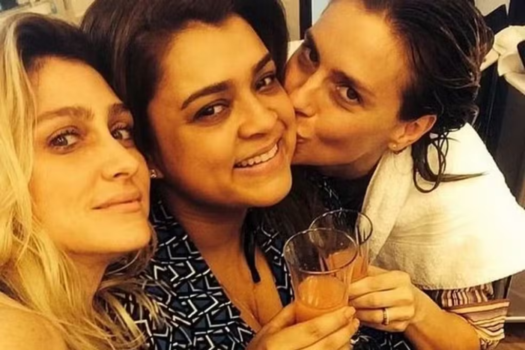 Amora Mautner, Preta Gil e Carolina Dieckmann sorriem juntas segurando copos com bebida, enquanto Carolina beija a bochecha de Preta Gil.