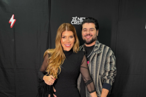 Paula Vaccari e o cantor Cristiano, da dupla Zé Neto e Cristiano, posam sorridentes em evento. Ela usa vestido preto e ele jaqueta estampada cinza e preta.