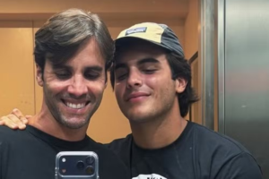 Daniel Cady sorri amplamente e seu filho Marcelo Sangalo, de olhos fechados e usando boné bege e cinza, posam para uma selfie refletida no espelho de um elevador residencial.