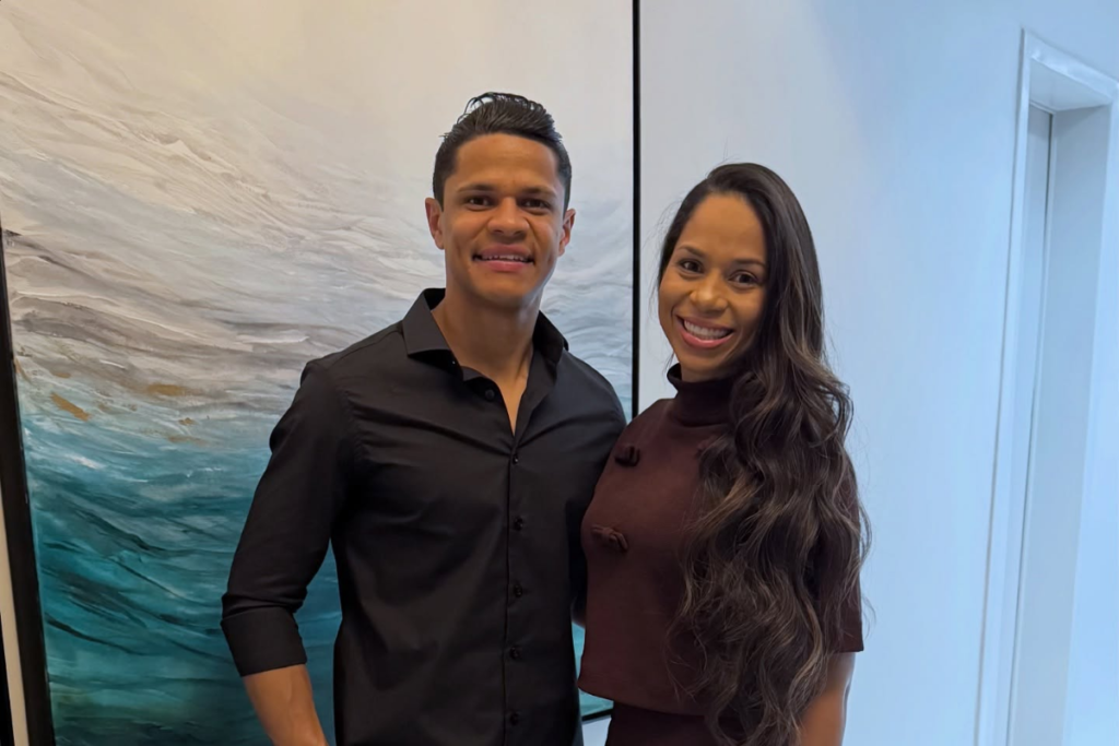 O jogador de futebol Douglas Santos posa sorridente ao lado de sua esposa, Cristiane Santos, em uma sala de estar. Ele veste uma camisa preta e ela um vestido marrom, em frente a uma grande pintura abstrata azul e branca.