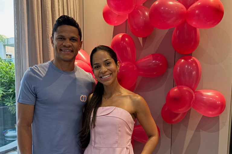 O jogador de futebol Douglas Santos posa sorridente ao lado de sua esposa, Cristiane Santos, em um ambiente decorado com balões cor-de-rosa.