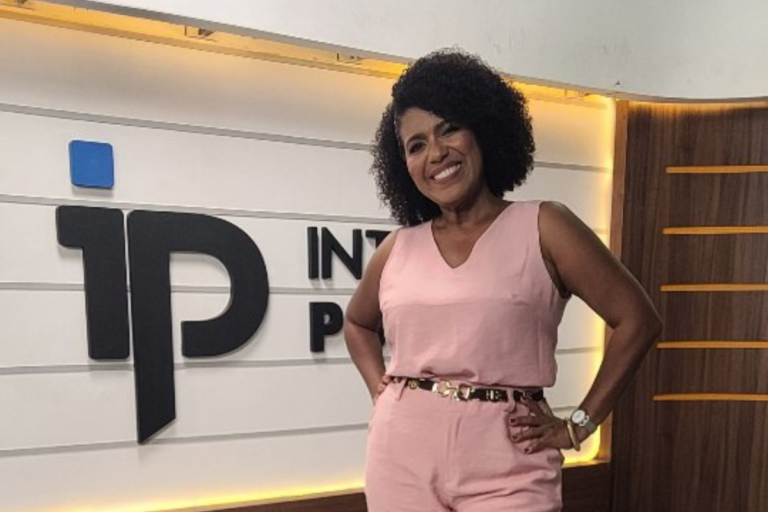 A jornalista Érika Leal posa sorridente para a câmera em um estúdio de televisão. Ela é uma mulher negra com cabelo cacheado volumoso e veste um macacão rosa claro sem mangas e com um cinto dourado. Ela está de pé, com as mãos na cintura, em frente a um painel branco com o logotipo '1P INTELLIGENCE PORTAL' em preto e azul, e detalhes em madeira.
