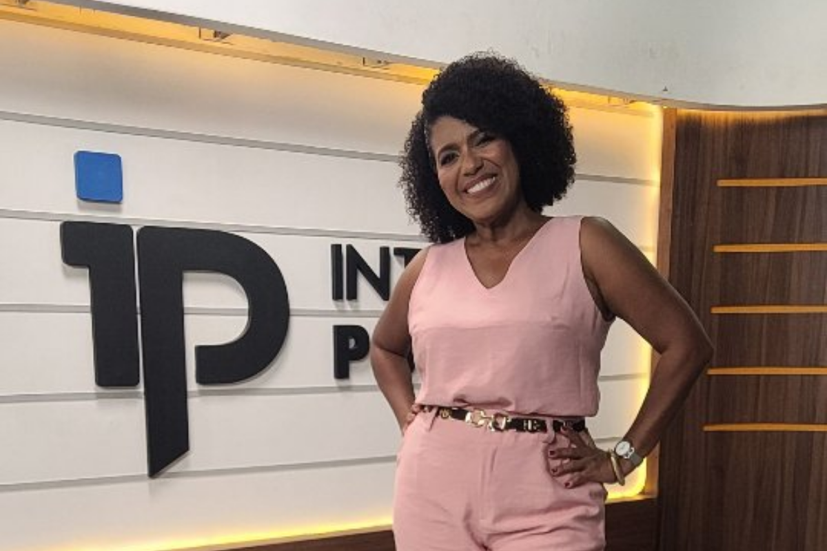 A jornalista Érika Leal posa sorridente para a câmera em um estúdio de televisão. Ela é uma mulher negra com cabelo cacheado volumoso e veste um macacão rosa claro sem mangas e com um cinto dourado. Ela está de pé, com as mãos na cintura, em frente a um painel branco com o logotipo '1P INTELLIGENCE PORTAL' em preto e azul, e detalhes em madeira.