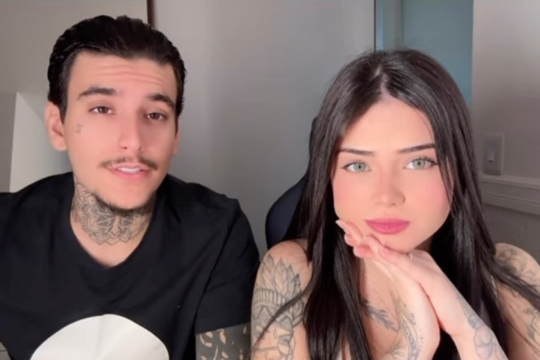 Arthur Urach e sua namorada, Maya, posam juntos para uma foto em plano médio. Arthur, à esquerda, tem tatuagens no rosto e pescoço e veste uma camiseta preta. Maya, à direita, tem longos cabelos escuros, olhos claros, braços tatuados e apoia o rosto nas mãos. Ambos olham para a câmera.