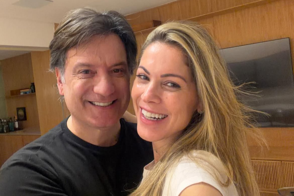 A jornalista Jacqueline Brazil e o narrador esportivo Luiz Carlos Jr. sorriem lado a lado em um registro descontraído dentro de um ambiente residencial.