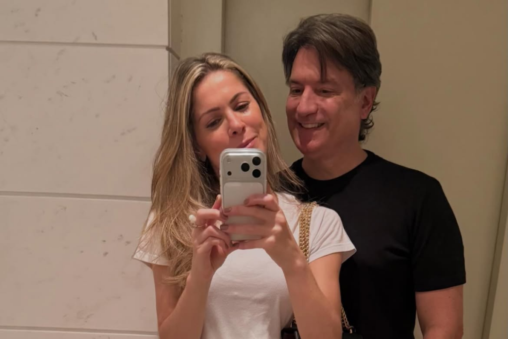 A jornalista Jacqueline Brazil segura um smartphone para tirar uma selfie no espelho ao lado do narrador esportivo Luiz Carlos Jr., que sorri enquanto olha para a tela do celular.
