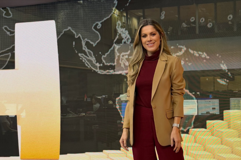 A jornalista Jacqueline Brazil sorri para a câmera em um estúdio de TV, vestindo um blazer bege e um conjunto bordô. Atrás dela, há um telão com um mapa-múndi estilizado e luzes de estúdio.