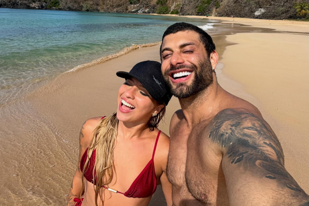 Jesus Luz e Rafaela Mesquita sorridentes durante um dia de sol em uma praia paradisíaca.