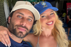 Jesus Luz e Rafaela Mesquita posam juntos e sorridentes, usando bonés, em um ambiente de celebração com decoração temática do Brasil.