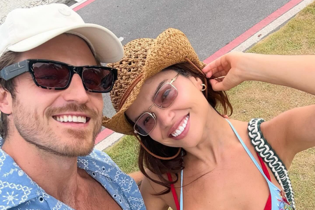 Juliette Freire sorri ao lado do noivo, Kaique Cerveny, em uma selfie ao ar livre perto de uma via pública. Ela usa chapéu de palha estilo cowboy, óculos de sol de lentes claras e biquíni azul com alça vermelha, enquanto ele veste camisa azul estampada, boné claro e óculos escuros.
