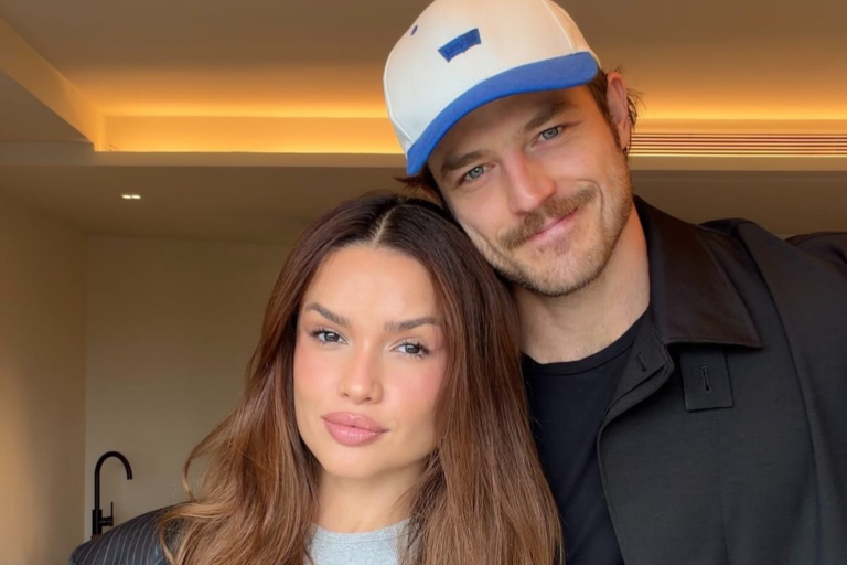 Juliette Freire e seu noivo, Kaique Cerveny, posam juntos sorrindo em ambiente interno com iluminação quente. Ela tem cabelos longos e castanhos, e ele usa um boné branco com aba azul e jaqueta preta.