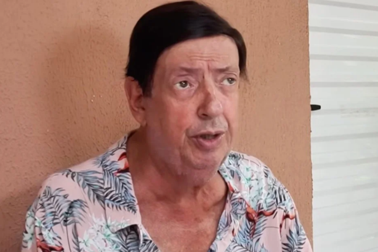 O ator Marcos Oliveira, conhecido pelo personagem Beiçola, posando para foto em plano médio com camisa estampada de flores e fundo de parede lisa.