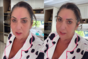 Margareth Serrão, de cabelos presos e vestindo um pijama branco com estampa de bolinhas pretas e detalhes em rosa, fala em um vídeo selfie gravado em uma cozinha moderna e iluminada ao fundo.