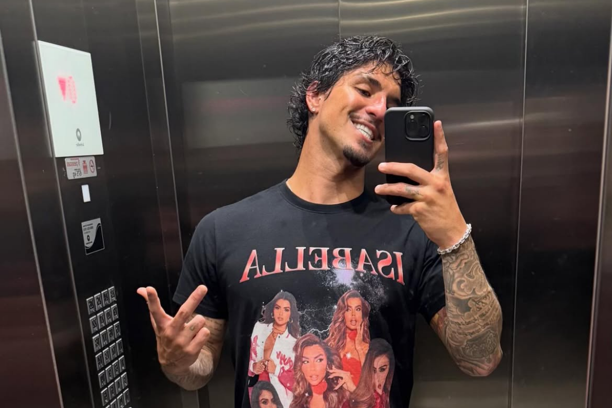 Gabriel Medina sorrindo e fazendo gesto com a mão enquanto tira selfie no espelho de um elevador, vestindo camiseta preta estampada.