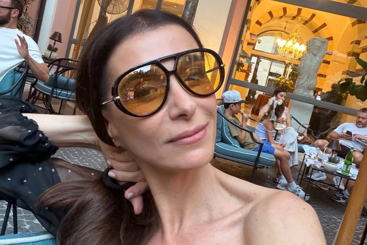 Mônica Martelli posa para selfie em ambiente externo com óculos escuros grandes de lente âmbar e blusa preta com poás.