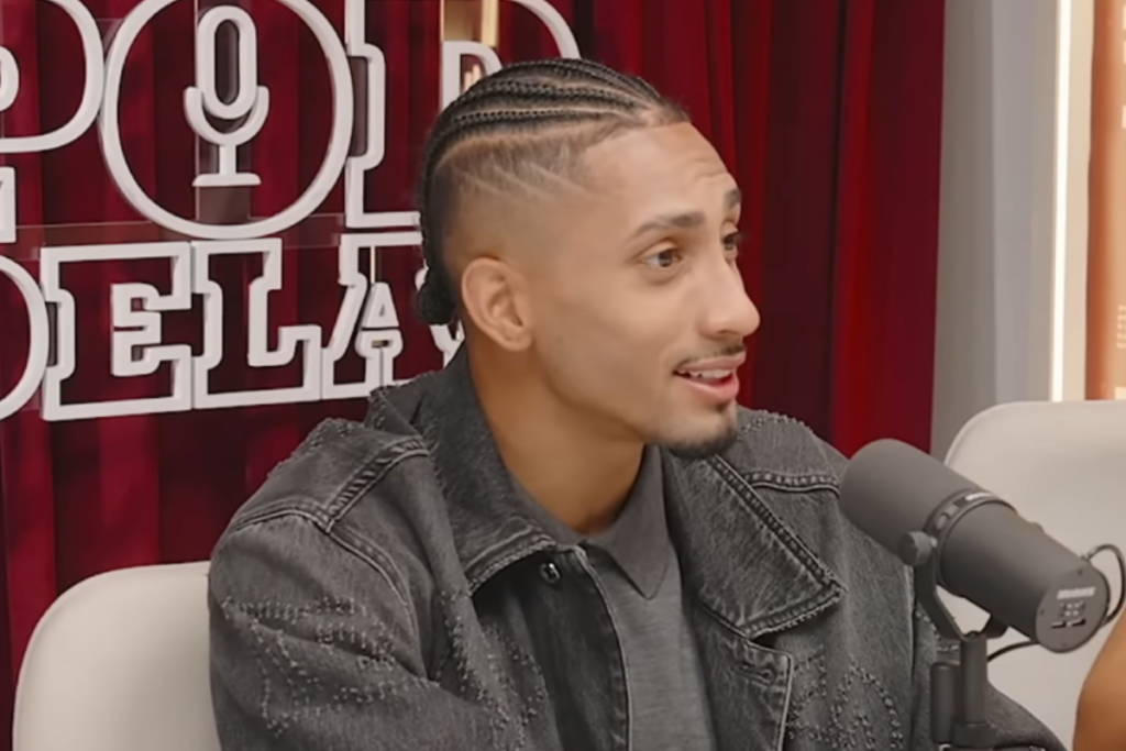 Raphinha com cabelo trançado, usando jaqueta jeans escura, participando de entrevista em podcast com microfone à frente.