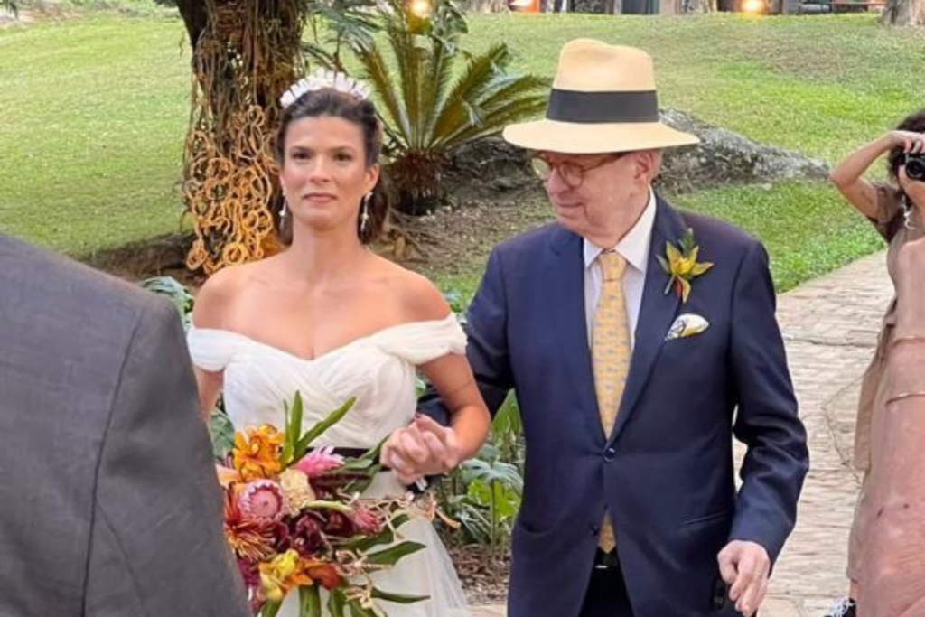 A noiva Maria Eduarda, usando um vestido branco ombro a ombro e tiara, caminha de braços dados com seu pai, o jornalista Renato Machado, que veste terno azul marinho, gravata amarela e um chapéu Panamá bege, em uma cerimônia de casamento ao ar livre.
