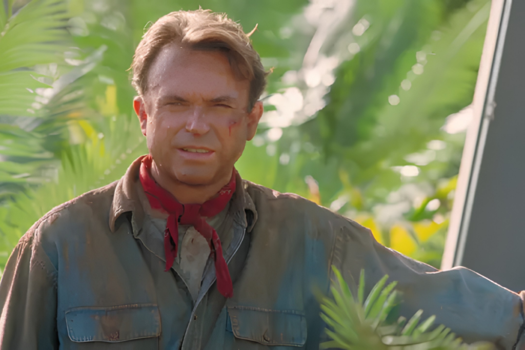 Ator Sam Neill como Dr. Alan Grant, sujo de lama e com lenço vermelho no pescoço, olhando para a câmera com expressão cansada e olhos semicerrados. Fundo desfocado com folhagem verde tropical e luz solar intensa.