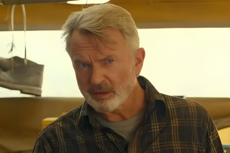 Sam Neill, ator neozelandês com cabelo grisalho e barba branca, vestindo uma camisa de flanela xadrez verde e marrom sobre uma camiseta cinza, olhando atentamente para a câmera com uma expressão séria. O fundo é desfocado, mostrando um ambiente de oficina com ferramentas e luz dourada.