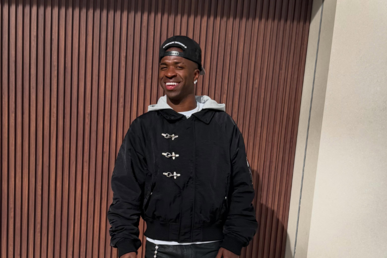 Vinicius Junior sorri em pé em frente a um painel de ripas de madeira vertical marrom. Ele veste uma jaqueta preta de estilo bomber com fechos metálicos frontais e capuz cinza claro, além de usar um boné preto virado para trás.