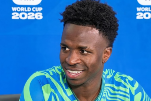 Vini Jr. sorrindo durante entrevista coletiva com fundo azul contendo a logomarca da Copa do Mundo 2026.