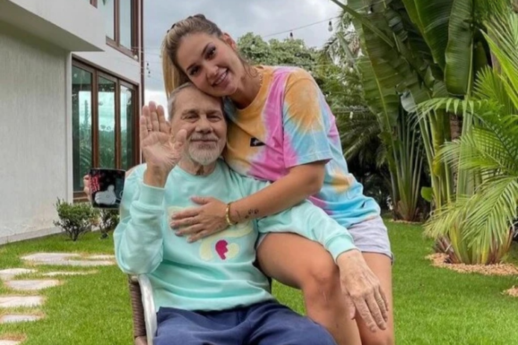 Virginia Fonseca abraça seu pai, Mário Ferrão, em um jardim gramado. Ela veste uma camiseta tie-dye colorida e sorri com o rosto encostado no dele. Mário, que está sentado, usa um moletom verde-água, segura uma caneca preta com a mão esquerda e acena para a câmera com a mão direita. Ao fundo, vê-se a fachada de uma casa moderna com portas de vidro e plantas de folhas largas.