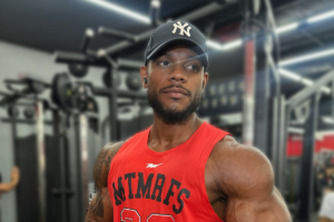 Fisiculturista Mailson Araújo Santos com físico musculoso em primeiro plano, vestindo regata vermelha e boné preto, posicionado dentro de uma academia com equipamentos de musculação desfocados ao fundo.