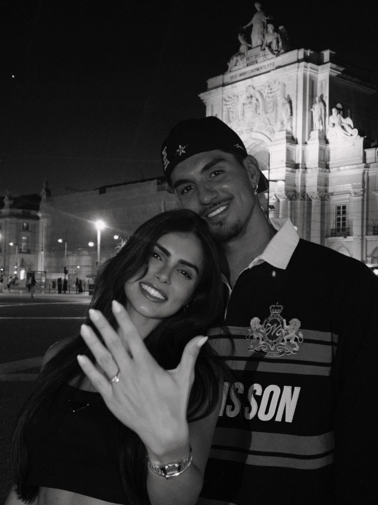 Isabella Arantes sorrindo e mostrando anel de noivado para a câmera enquanto é abraçada por Gabriel Medina, em frente a um monumento histórico iluminado à noite.