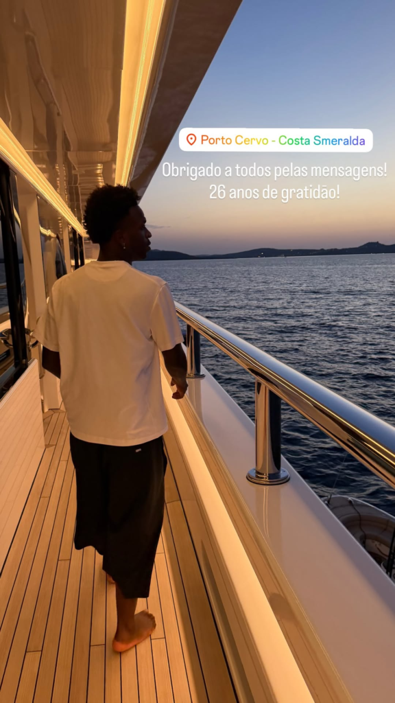 Vinicius Junior, jogador de futebol, de costas a bordo de um iate de luxo durante o entardecer. Ele veste camiseta branca e bermuda preta, caminhando descalço pelo convés de madeira enquanto observa o mar de Porto Cervo, na Itália. Há um texto sobreposto na imagem celebrando seu aniversário de 26 anos.