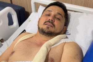 Ricardo Castella, primo da cantora Ana Castela, deitado em uma maca de hospital com os olhos semifechados. Ele está sem camisa, sob lençóis brancos, e utiliza uma tipoia ortopédica bege que passa por seus ombros e peito.