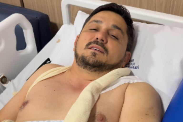 Ricardo Castella, primo da cantora Ana Castela, deitado em uma maca de hospital com os olhos semifechados. Ele está sem camisa, sob lençóis brancos, e utiliza uma tipoia ortopédica bege que passa por seus ombros e peito.