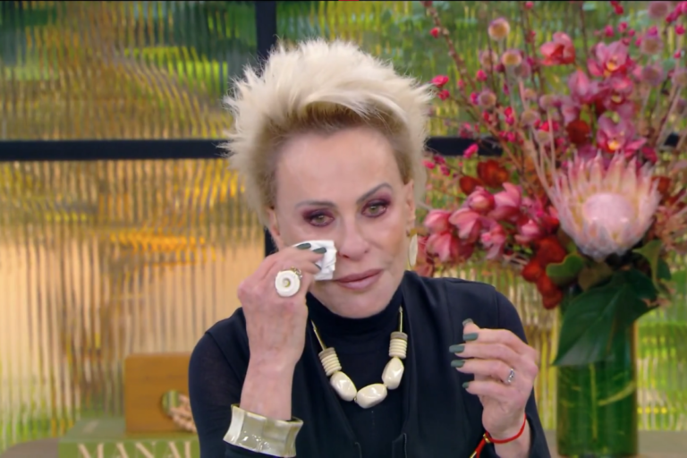A apresentadora Ana Maria Braga, com seu característico cabelo loiro espetado, vestindo roupa preta e colar de contas, enxuga uma lágrima do rosto com um lenço branco durante uma transmissão ao vivo.