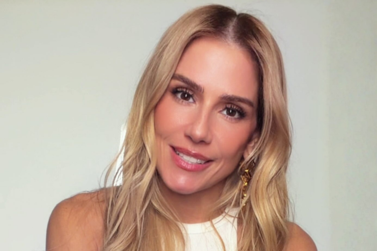 Deborah Secco de cabelos loiros ondulados, sorrindo levemente para a câmera, vestindo blusa branca e brincos dourados longos.