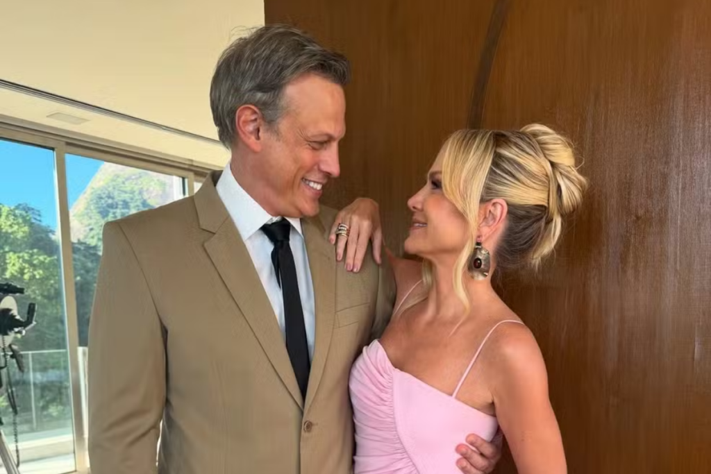 A apresentadora Eliana e seu marido, o diretor de TV Adriano Ricco, posam sorrindo e se olhando. Ela usa um vestido longo rosa com recortes e ele um terno bege claro. Ao fundo, vê-se a paisagem através de uma janela de vidro.