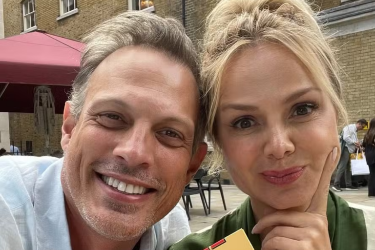 A apresentadora Eliana e seu marido, o diretor Adriano Ricco, sorriem juntos em uma selfie tirada ao ar livre em uma área externa com edificações e guarda-sol ao fundo.
