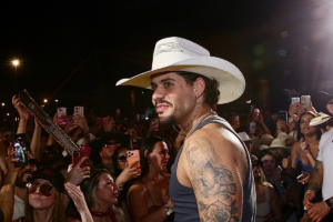 Zé Felipe de perfil, sorrindo levemente durante um evento noturno. Ele usa um chapéu de caubói branco, brincos de cruz e uma regata cinza, deixando em evidência as tatuagens de Jesus Cristo e rosas no braço direito. Ao fundo, uma multidão de fãs levanta celulares para fotografá-lo.