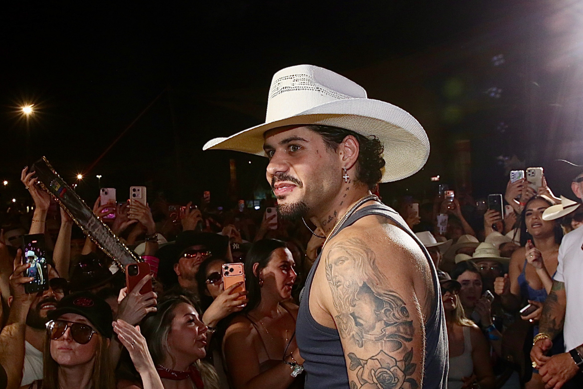 Zé Felipe de perfil, sorrindo levemente durante um evento noturno. Ele usa um chapéu de caubói branco, brincos de cruz e uma regata cinza, deixando em evidência as tatuagens de Jesus Cristo e rosas no braço direito. Ao fundo, uma multidão de fãs levanta celulares para fotografá-lo.