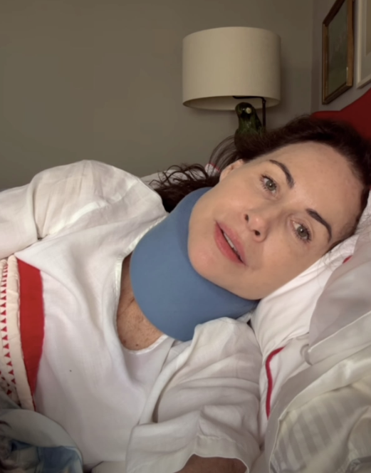 A atriz Minnie Driver deitada na cama usando um colar cervical azul no pescoço