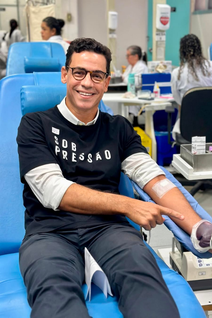 Márcio Maranhão sentado em poltrona de coleta, sorrindo e apontando para curativo no braço após doar sangue em ambiente hospitalar, vestindo camiseta "SOB PRESSÃO".