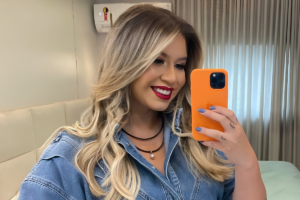 Marilia Mendonça sorrindo para a câmera enquanto segura um celular laranja para uma selfie. Ela usa uma jaqueta jeans azul-clara, um colar de miçangas escuras com pingente e tem cabelos loiros ondulados. A foto foi tirada em um ambiente interno com uma cama e cortinas ao fundo.