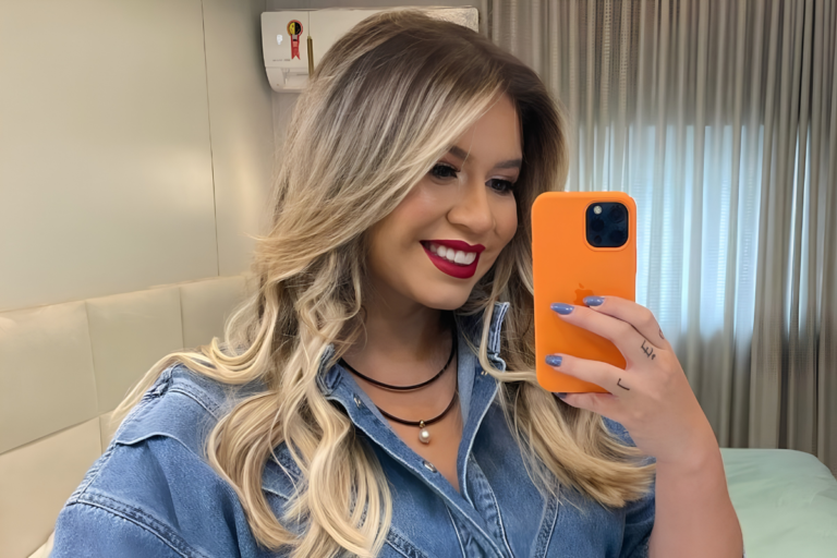 Marilia Mendonça sorrindo para a câmera enquanto segura um celular laranja para uma selfie. Ela usa uma jaqueta jeans azul-clara, um colar de miçangas escuras com pingente e tem cabelos loiros ondulados. A foto foi tirada em um ambiente interno com uma cama e cortinas ao fundo.